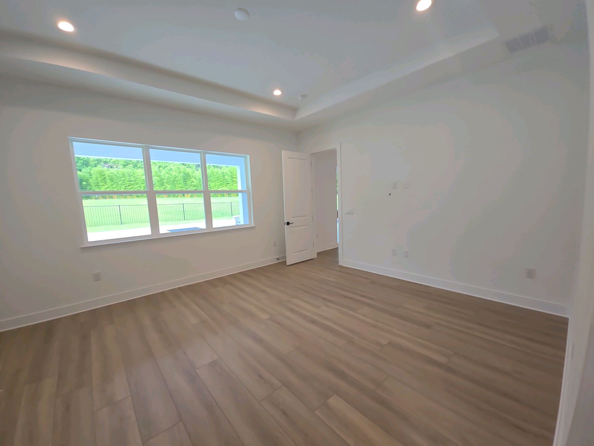 An empty finished bedroom with a tray ceiling, wood-look floor and a window onto the back yard, before closing