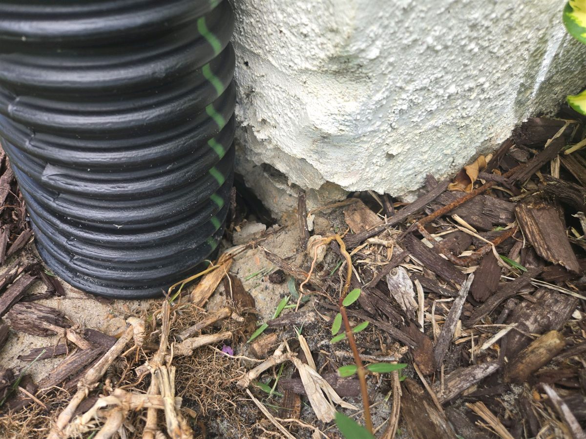 A corrugated downspout drain pipe against a stucco wall where the soil has settled and the stucco edge is exposed