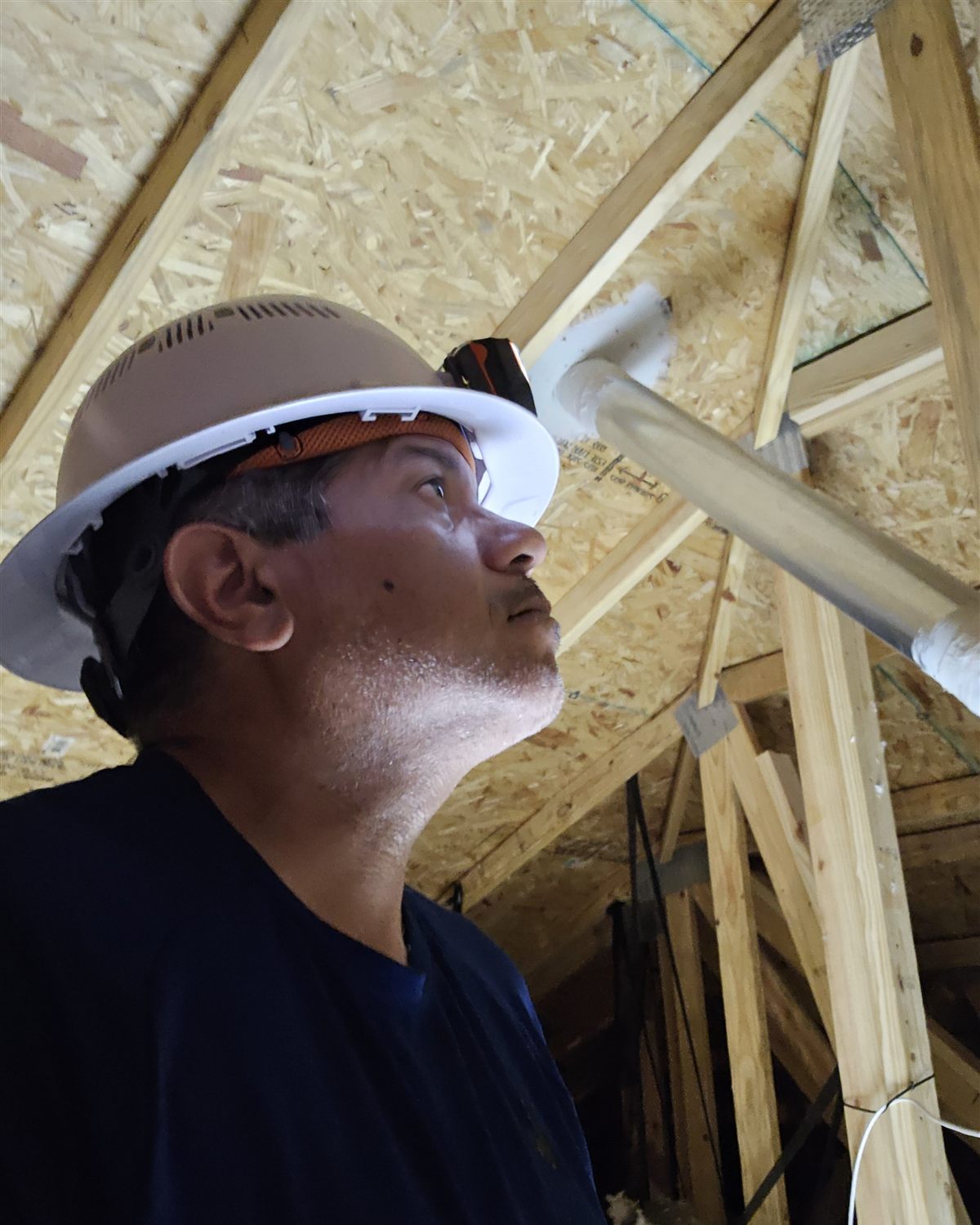 The Home IQ inspector in a hard hat, looking up at roof trusses and a duct run in an attic under construction