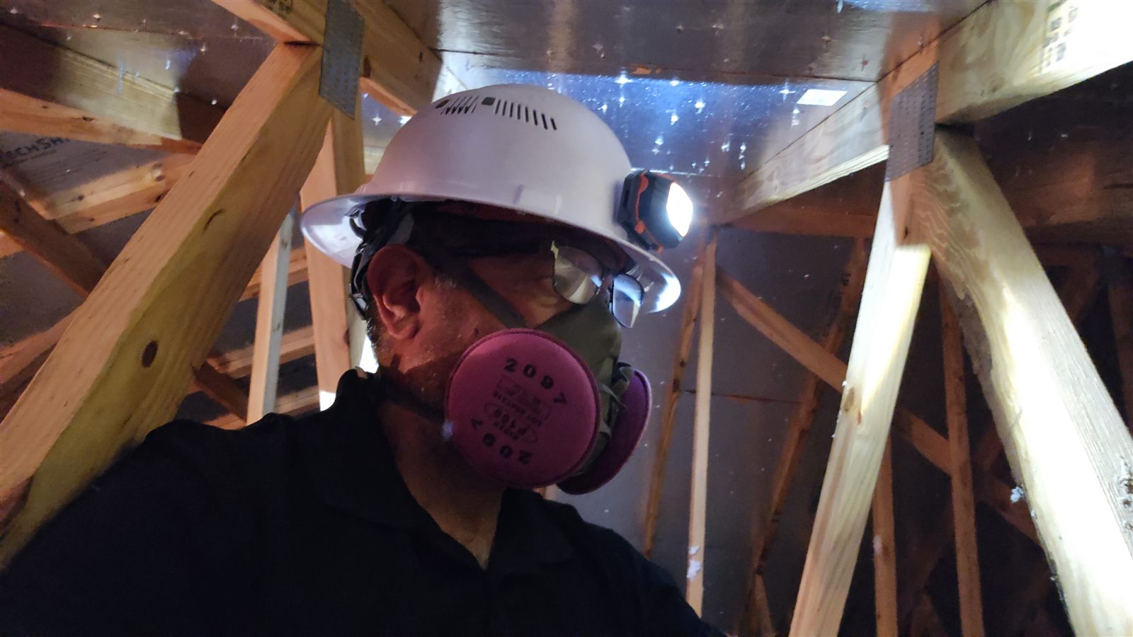 The Home IQ inspector in a hard hat and respirator looking up at trusses and ductwork in an attic