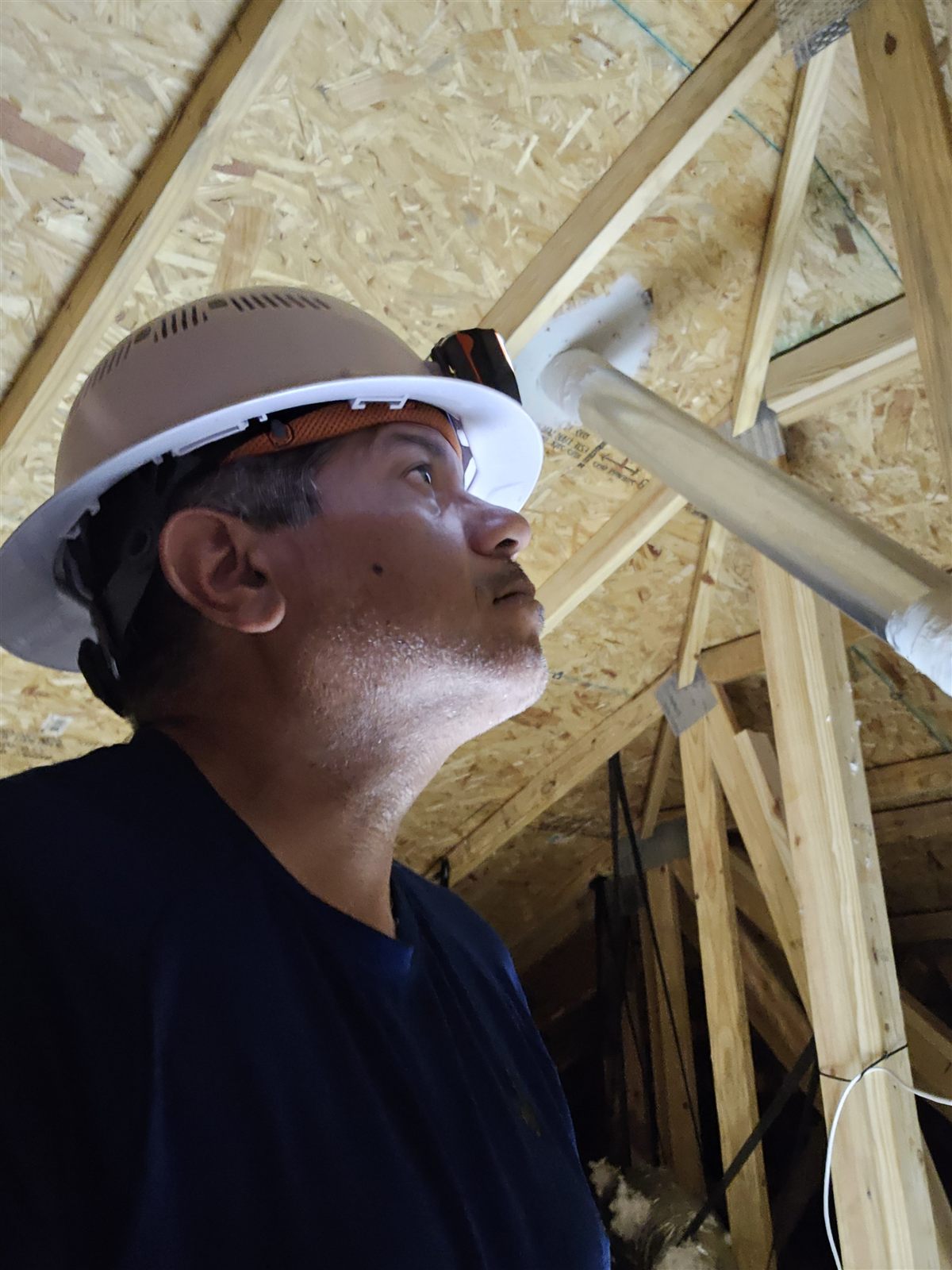 The Home IQ inspector in a hard hat, looking up at roof trusses and a duct run in an attic under construction