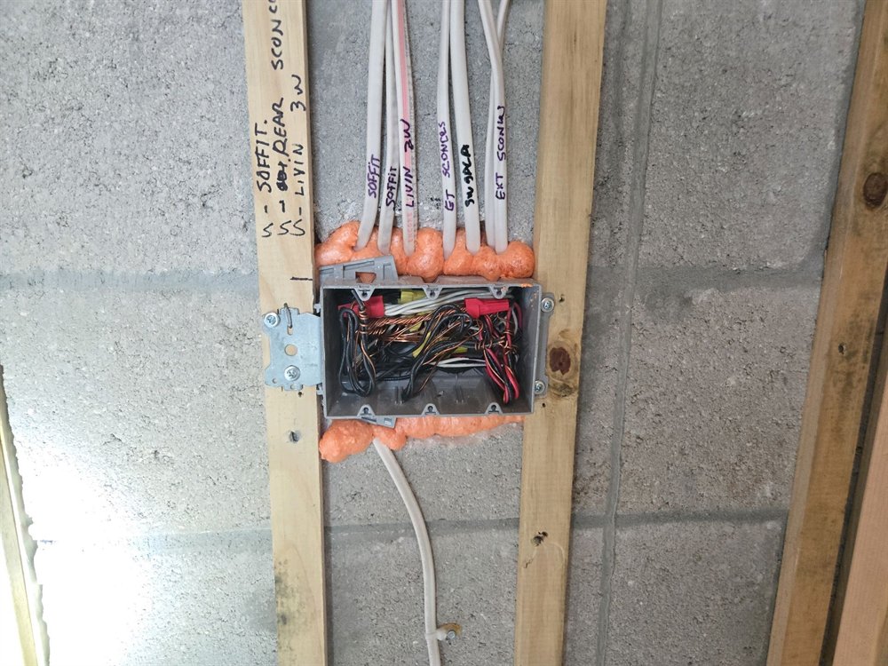 Open three-gang electrical box on a furring strip against a block wall, with cables entering through a foam-sealed penetration and connectors visible inside