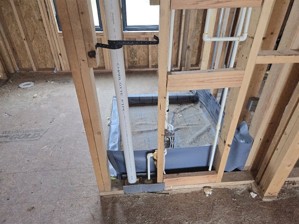 Shower rough-in in a framed wall, showing the drain stack strapped to a stud, the shower pan liner, supply lines and a steel nail plate at the base