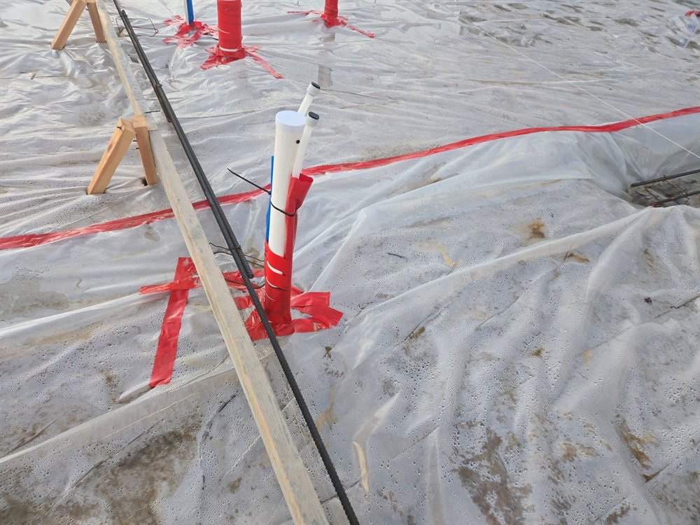 Capped plumbing stub-ups rising through the vapor barrier on a slab pad, each penetration wrapped and taped where the pipe passes through the sheeting